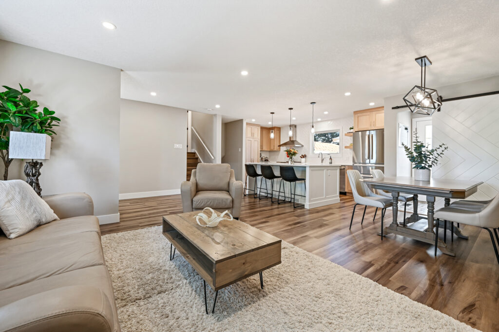 Open-concept living space with beige walls, wooden floors, a cozy sofa and armchair, modern coffee table, kitchen island with barstools, and a dining area with a rectangular table and contemporary lighting.
