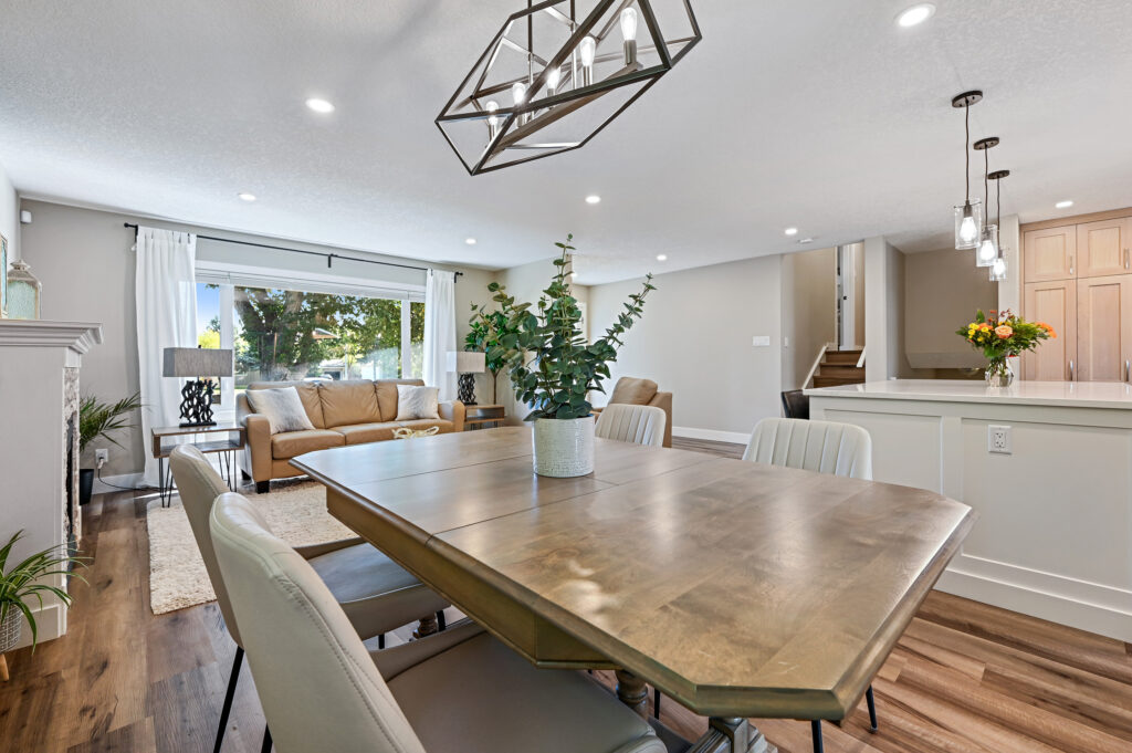 Modern open-concept living and dining area with a wooden table, beige chairs, couch, large windows with white curtains, hardwood floors, and pendant lights over the kitchen island.