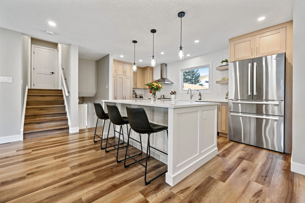 Modern kitchen with wood floors, a large island with three black barstools, pendant lights, stainless steel appliances, light wood cabinets, and a window above the sink. Stairs are visible to the left.
