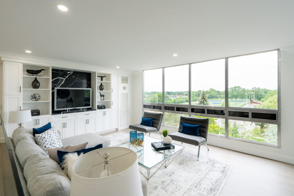 Bright, modern living room with white walls, large windows overlooking greenery, gray sofa, navy blue chairs, glass coffee table, built-in shelves, and a wall-mounted TV. Natural light fills the space.