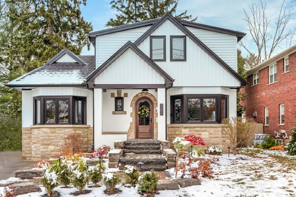 Two-story house with white siding, stone accents, and black trim, featuring a central brown door with a wreath, large bay windows, and a small front garden with patches of snow on the ground.