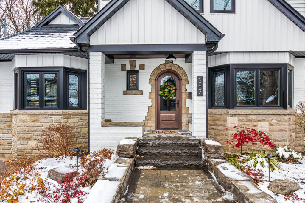 Front entrance of a house with stone steps, a brown door decorated with a wreath, large black-trimmed windows, and snow covering the ground and plants in the landscaping.