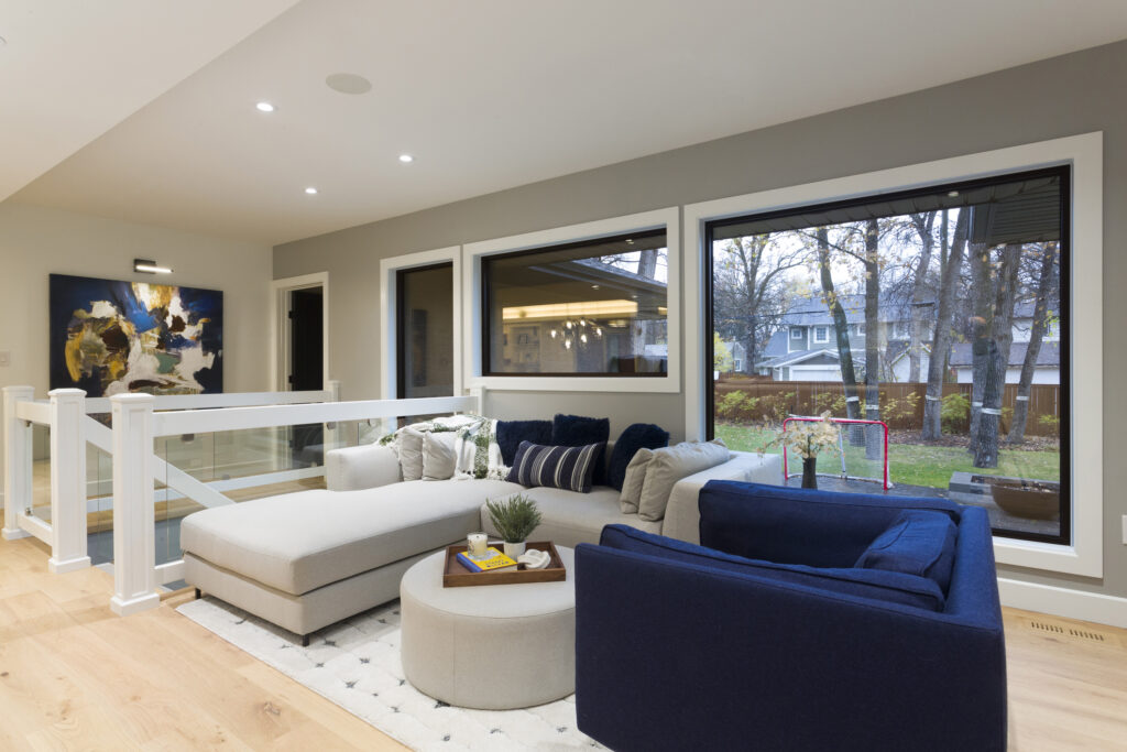 A modern living room with large windows, a sectional sofa in light and navy colors, a round ottoman, abstract wall art, and views of a backyard with trees and a hockey net.