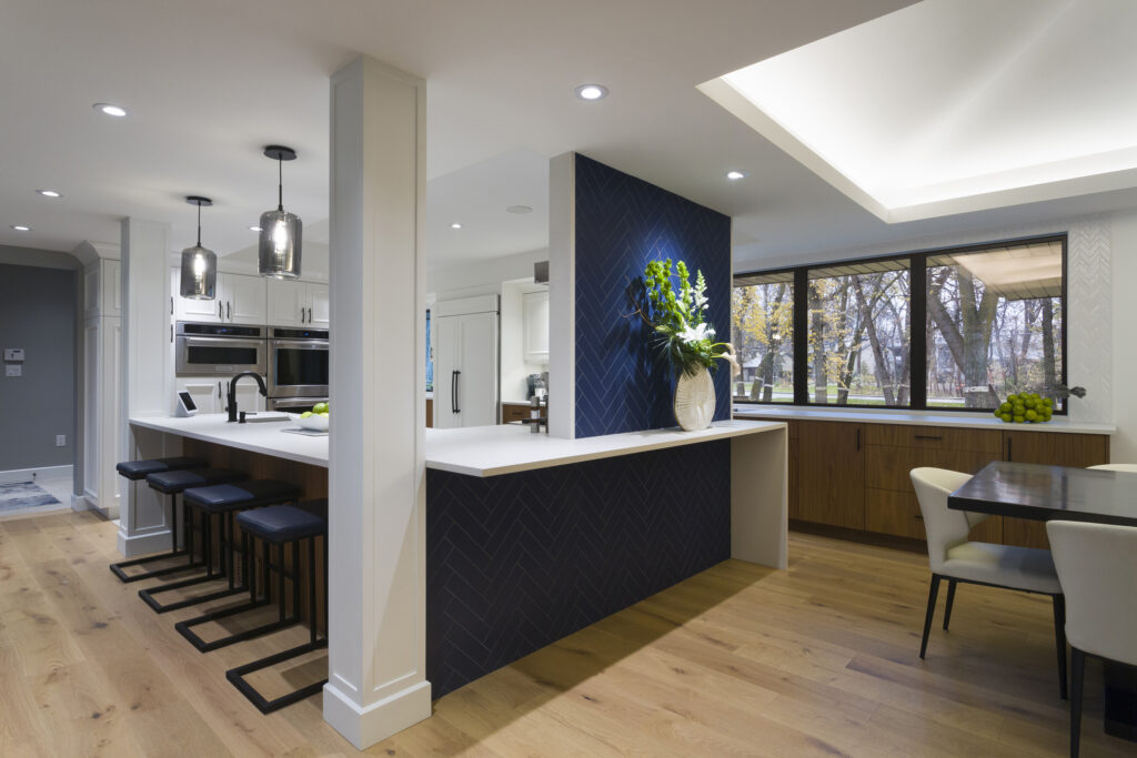 Modern kitchen with a white island, navy blue accent wall, black bar stools, pendant lights, wooden floor, large windows, and a dining area with white chairs and a wooden table. A vase with green plants sits on the counter.