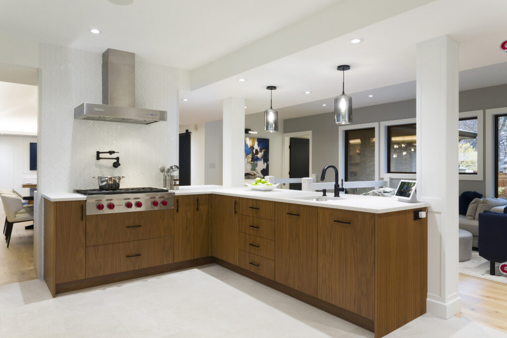 Modern kitchen with wood cabinets, white countertops, stainless steel stove, and range hood. Pendant lights hang above the counter, and an open layout connects to a living area with dark-framed windows.