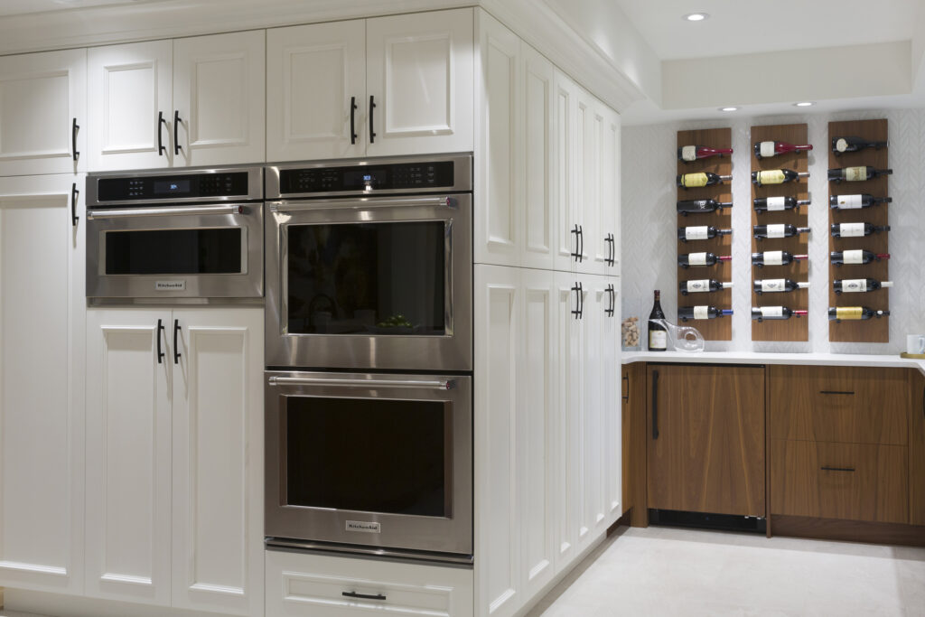 A modern kitchen with white cabinets, built-in stainless steel ovens, and a microwave. In the corner, there is a wooden wine rack mounted on the wall displaying bottles, above a brown cabinet and countertop.