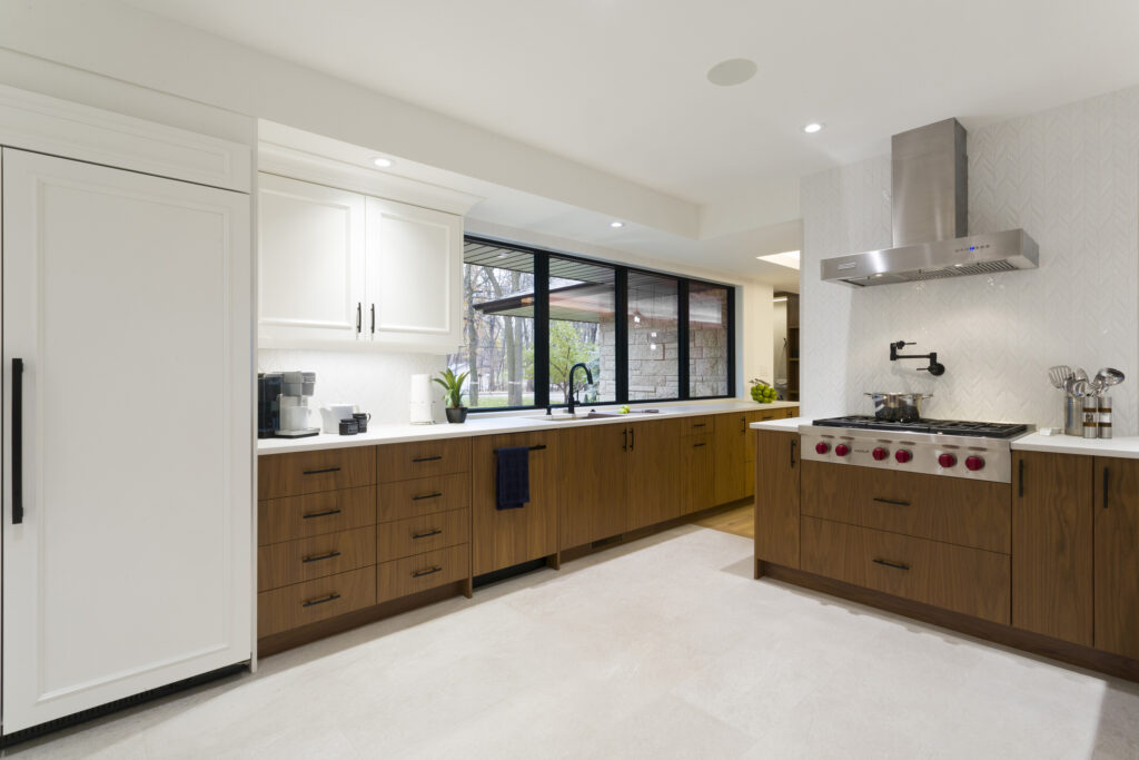 Modern kitchen with white and wood cabinets, a large white refrigerator, stainless steel appliances, a wide window above the sink, and light-colored floors. The space is bright and tidy with minimal decor.