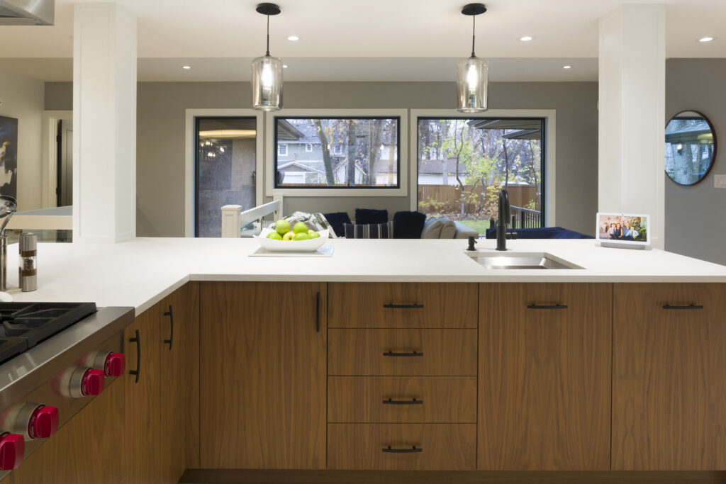 Modern kitchen with wood cabinets, white countertops, two pendant lights, a sink, and a fruit bowl. Large windows in the background show trees and houses outside. A photo frame sits on the counter near the windows.