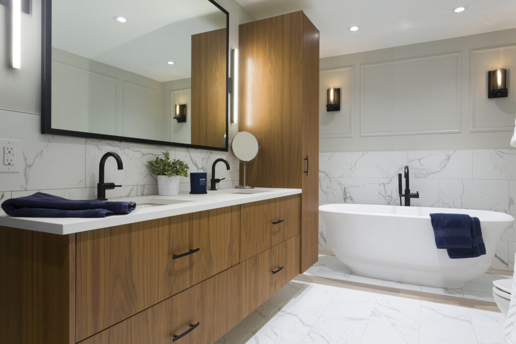 Modern bathroom with a double-sink wooden vanity, large mirror, black fixtures, white freestanding bathtub, marble tile floor and walls, wall sconces, and a potted plant on the counter.