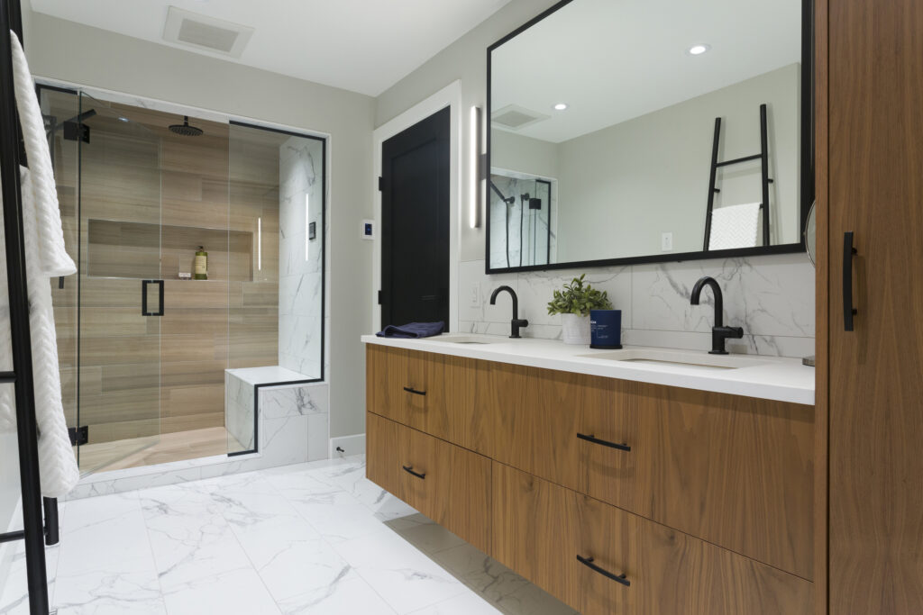 Modern bathroom with a double wooden vanity, two black faucets, a large mirror, and a glass-enclosed shower with a bench. The space features white marble floors and walls, and minimalist decor.
