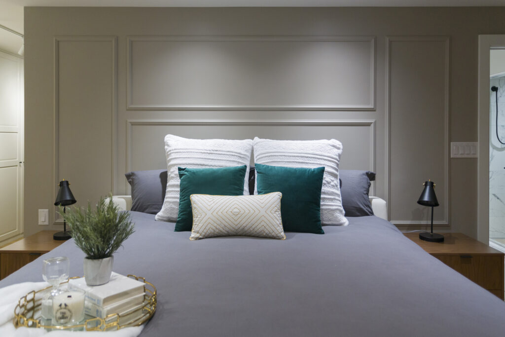 A neatly made bed with gray bedding, green and white pillows, and two black lamps on wooden bedside tables. A decorative tray with a plant, candle, and glass sits at the foot of the bed. A bathroom is visible on the right.