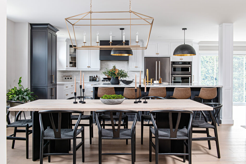 Modern kitchen and dining area with a large wooden table, black chairs, and a decorative centerpiece. The kitchen features white cabinets, a black island with brown stools, pendant lights, and stainless steel appliances.