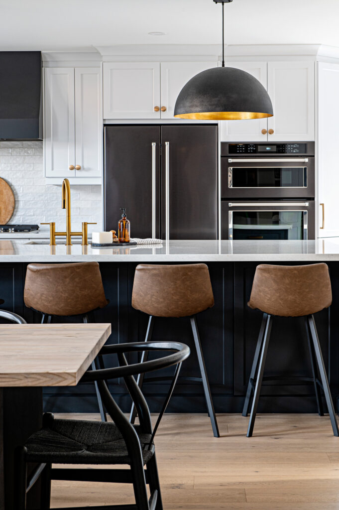 Modern kitchen with a large island, three brown barstools, gold fixtures, black and white cabinetry, stainless steel double ovens, and a black pendant light hanging above the island.
