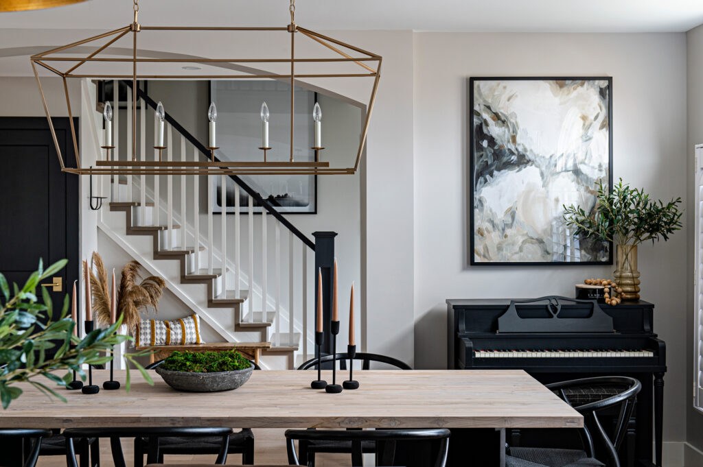 Modern dining room with a light wooden table, black chairs, and a gold geometric chandelier. A staircase and abstract art are in the background, with a black piano and green plants adding decorative touches.