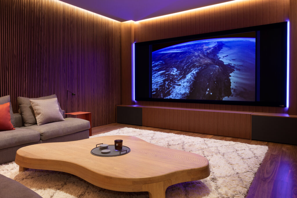 A modern home theater with a large screen displaying Earth from space, wood-paneled walls, a cozy sectional sofa with pillows, a wooden coffee table, and a plush white rug. Soft ambient lighting highlights the room.