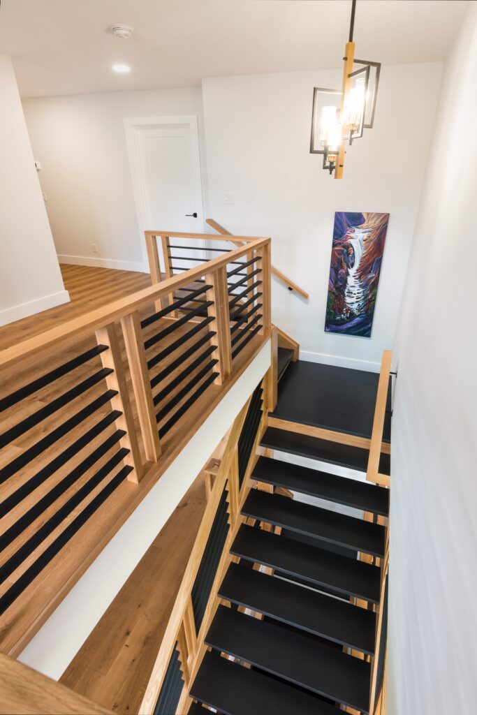 A modern indoor staircase with black steps, wooden railings, and horizontal black balusters, leading to a hallway. A painting of a tiger and a geometric pendant light hang on the white wall.