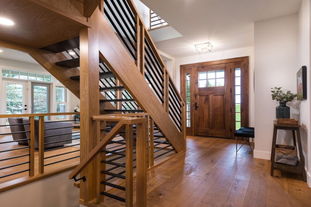 Modern home entryway with wooden floors, a wooden staircase with black metal railings, a wooden front door with glass panels, a bench, a small table with a plant, and light streaming in from windows.