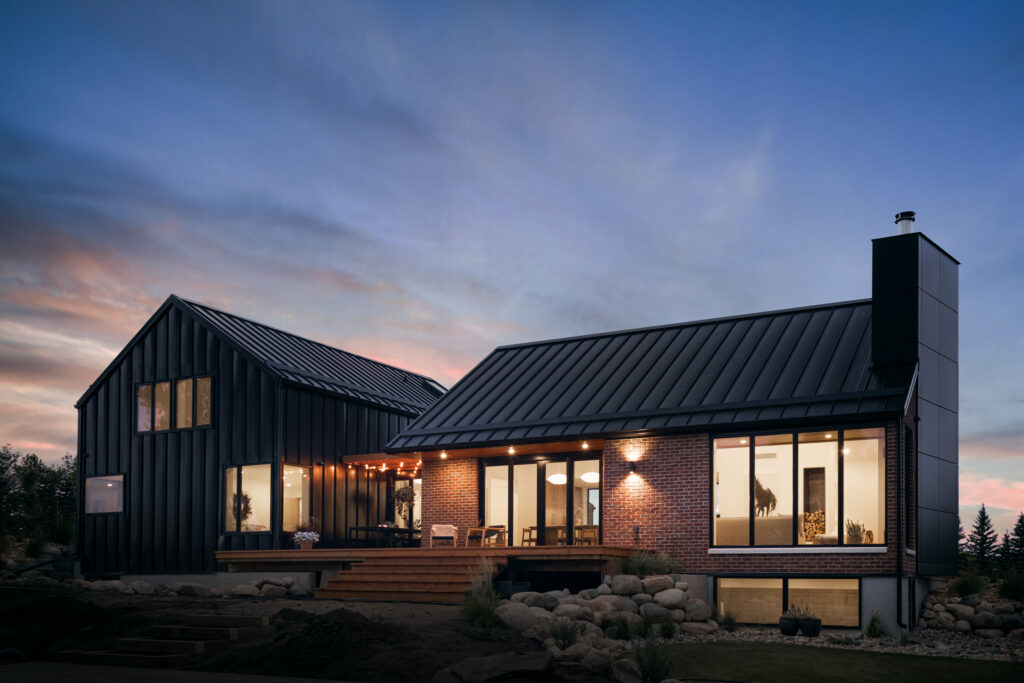 Modern house at dusk with black metal roofs, large windows, and brick walls. Warm interior and exterior lights are on, illuminating the patio with outdoor seating. The sky is partly cloudy with a soft pink and blue sunset.