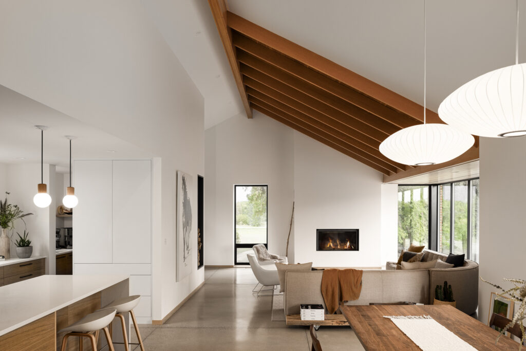 A modern open-concept living space with a sloped wooden ceiling, pendant lights, a fireplace, neutral-toned furniture, large windows, and a dining area with a wooden table and chairs.