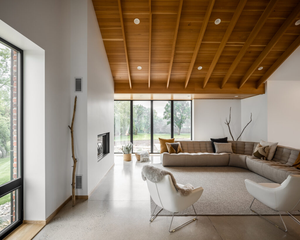 A modern living room with a wooden ceiling, large corner windows, a beige sectional sofa, white armchairs, and minimalist decor. Natural light fills the space, highlighting indoor plants and a view of greenery outside.