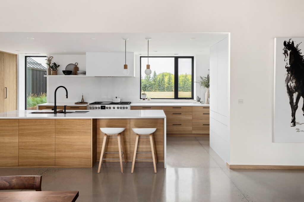 Modern kitchen with wooden cabinets, white countertops, two white bar stools, black sink faucet, and pendant lights. Large window offers a view of greenery; a horse painting hangs on the right wall. The space is bright and minimalist.