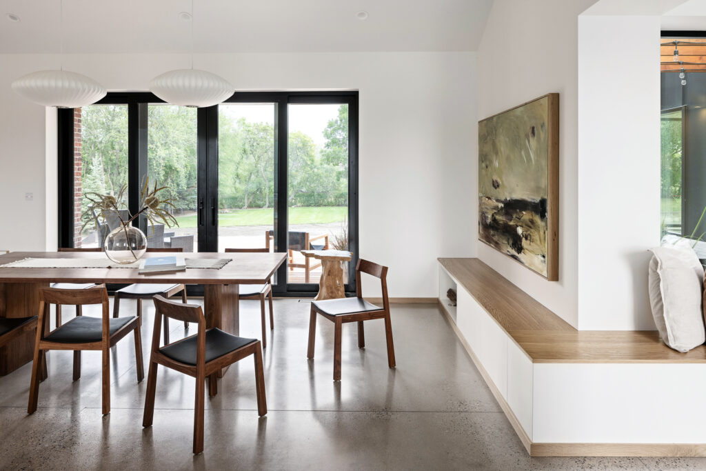Minimalist dining room with a wooden table, six wooden chairs, two pendant lights, large windows overlooking greenery, a bench with cushions, and abstract art on the wall.