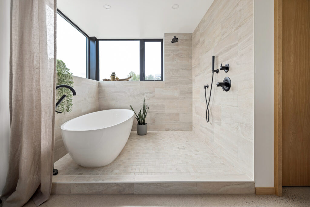 Modern bathroom with a freestanding white bathtub, black fixtures, a walk-in shower, large windows, beige tile walls and floor, a potted plant, and natural light streaming in.