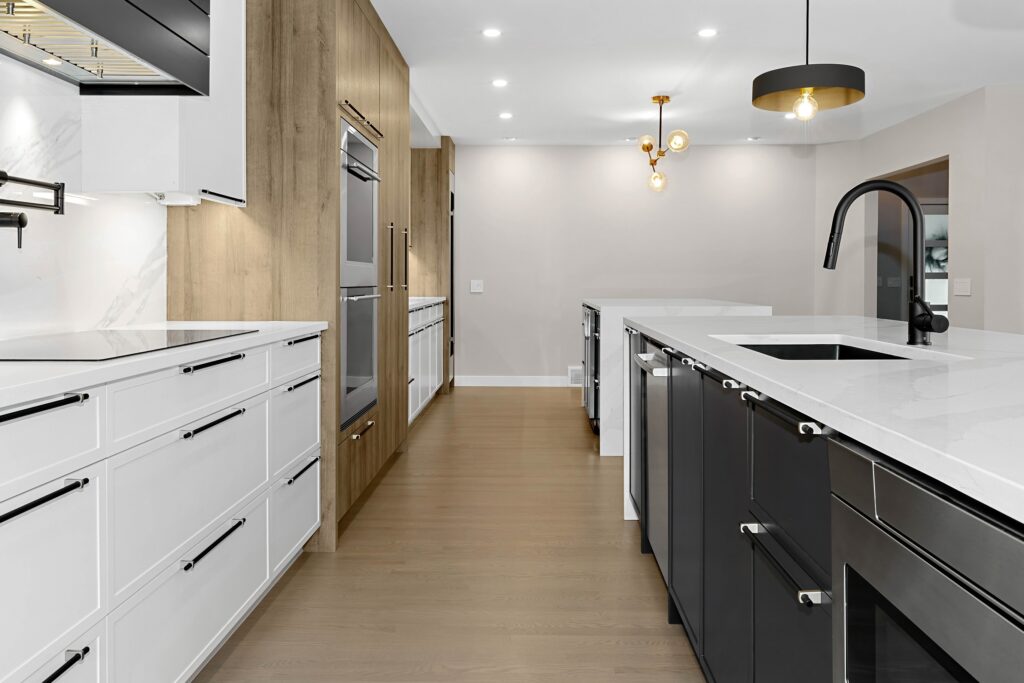 Modern kitchen with white and black cabinets, wood accent wall, built-in ovens, white countertops, black faucet, and pendant lighting. The space features a clean, minimalist design with recessed ceiling lights.