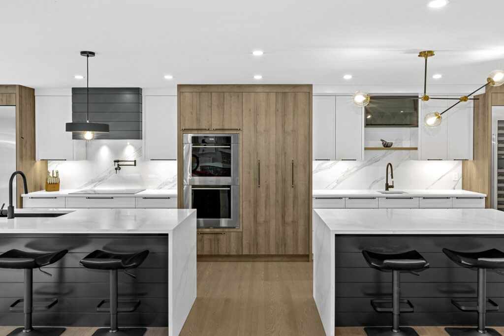 Modern kitchen with white cabinets, wood accents, a built-in oven, two islands with black stools, marble countertops, pendant lights, and a wooden floor. The space is bright and sleek with gold and black fixtures.