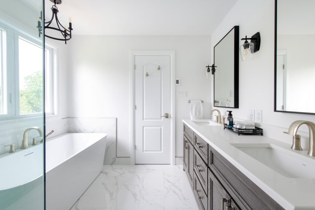 Bright, modern bathroom with a freestanding white bathtub, double vanity with dark cabinets and two sinks, large mirrors, marble floor tiles, and a central white door. Natural light pours in from a large window.