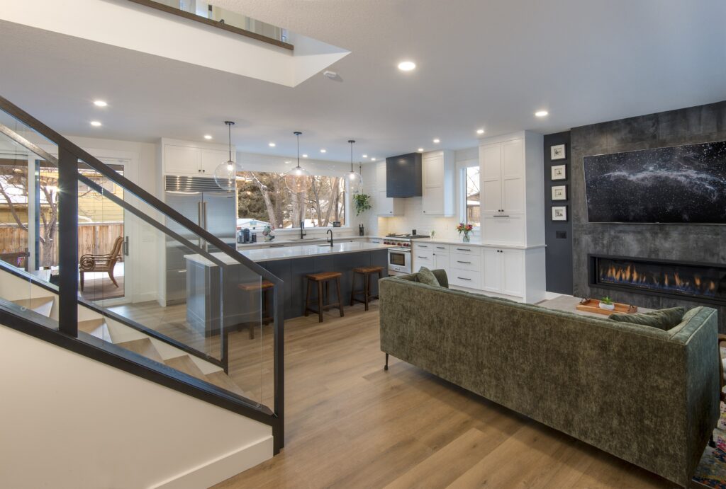 Modern open-concept living space with a green sofa, glass-railing staircase, large kitchen with island and pendant lights, and a dark accent wall with a fireplace and mounted TV. Large windows provide natural light.