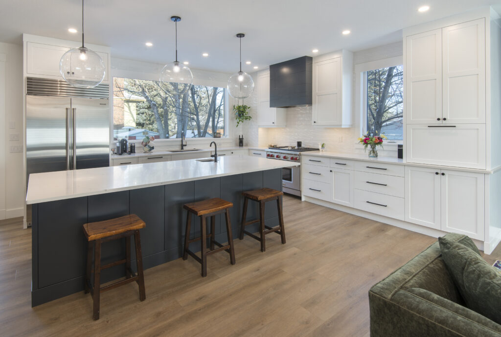 Modern kitchen with white cabinets, a large island with a white countertop, three wooden stools, stainless steel appliances, pendant lights, and large windows letting in natural light.