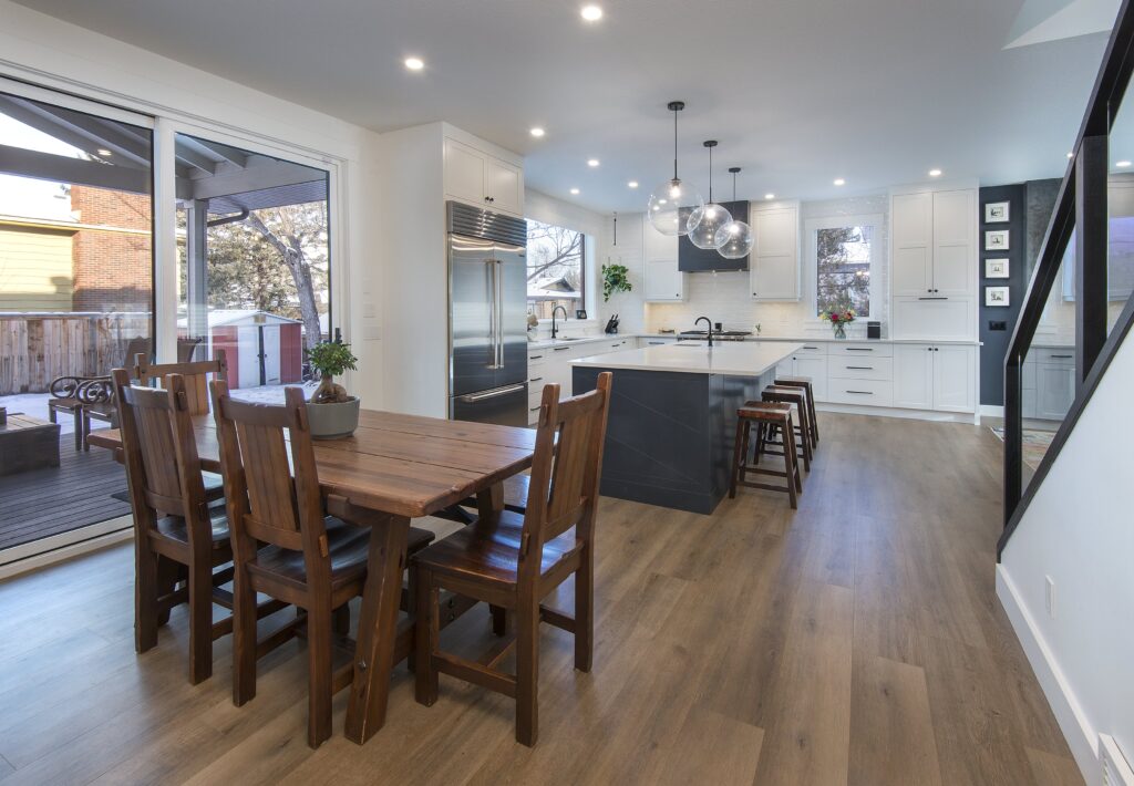 Modern kitchen and dining area with wood floors, wooden dining table and chairs, a large island with stools, white cabinets, pendant lights, and sliding glass doors leading to an outdoor patio.