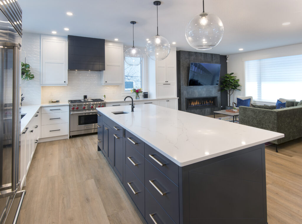 Modern kitchen with a large white island, navy blue cabinets, stainless steel appliances, pendant lights, a fireplace, and a living area with a green sofa. Light wood flooring and large windows create a bright, open space.