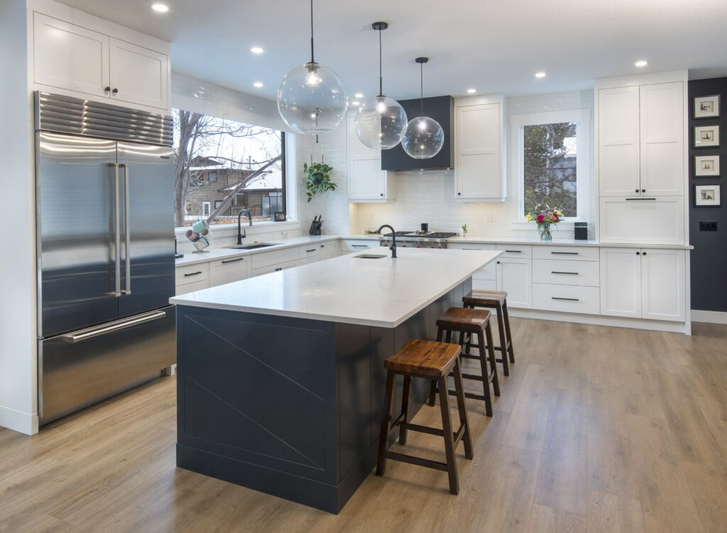 Modern kitchen with white cabinets, stainless steel appliances, a large island with a white countertop, dark blue base, three wooden stools, globe pendant lights, and light wood flooring.