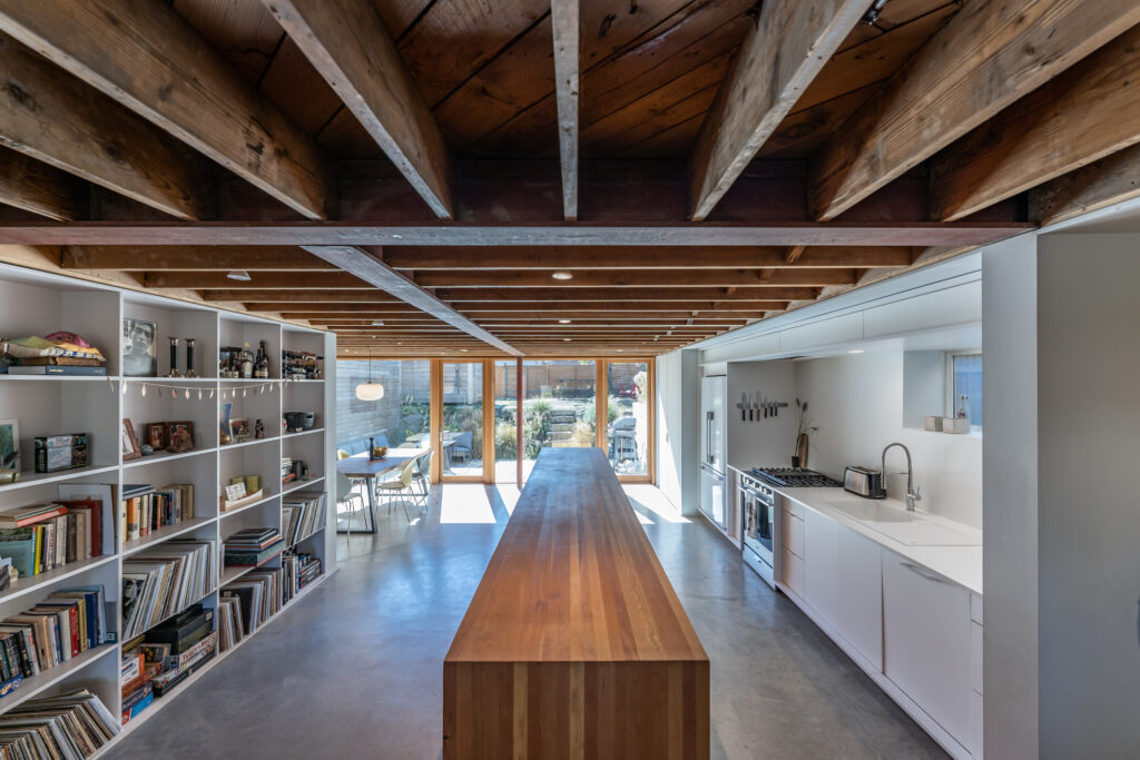 Modern kitchen with a long wooden island, open shelves filled with books and decor, exposed wooden ceiling beams, white cabinets, and large windows overlooking a patio and garden.