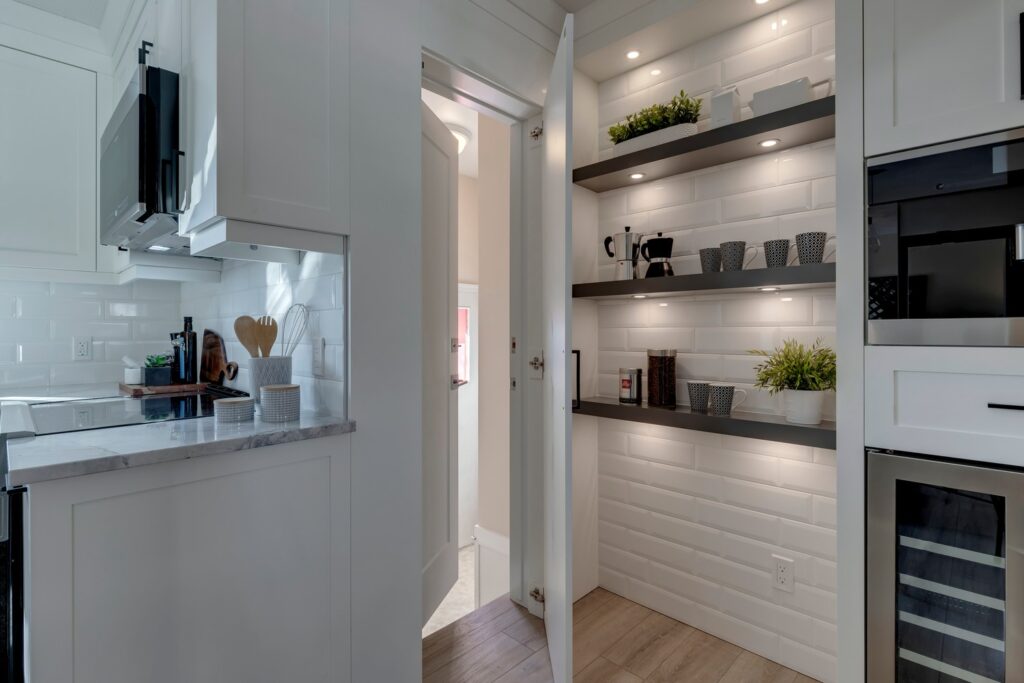 A modern kitchen with a white interior featuring an open pantry with shelves holding dishes, mugs, storage jars, and potted plants. The kitchen has marble countertops, white cabinets, and built-in appliances.