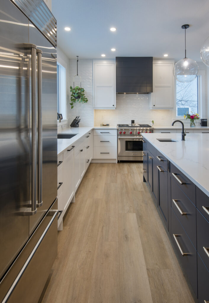 Modern kitchen with stainless steel refrigerator, white cabinets, dark island cabinets, wood flooring, pendant lights, a gas stove, and large windows providing natural light. A potted plant sits on the counter.