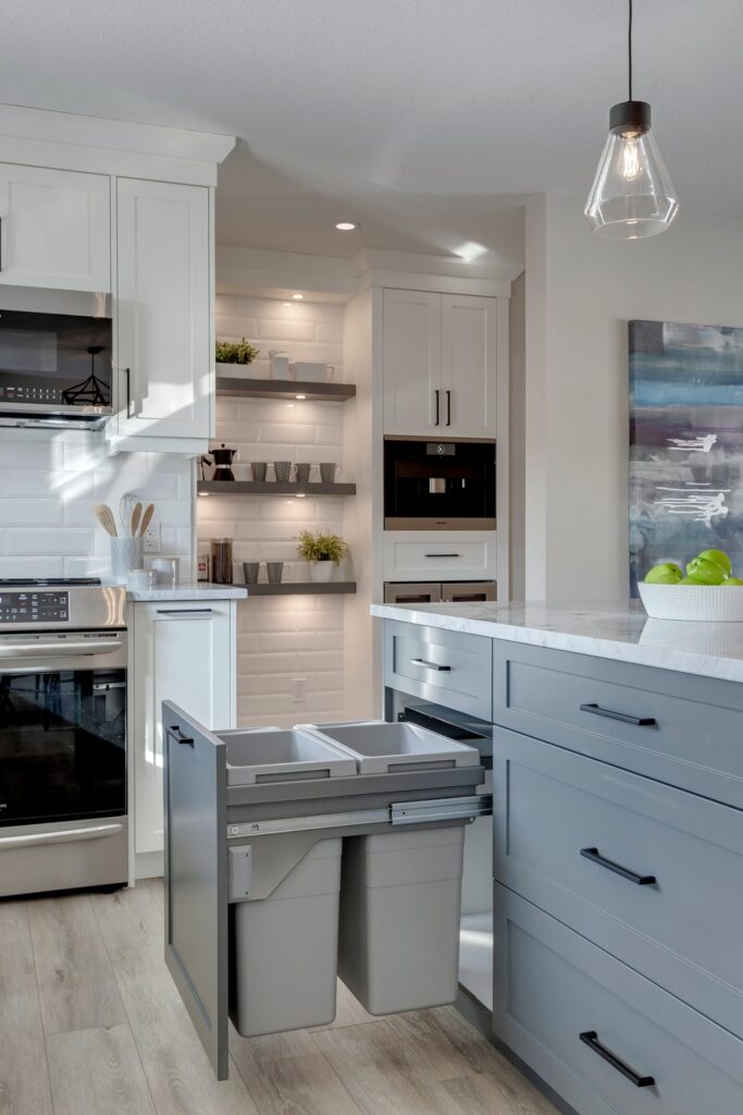 Modern kitchen with white cabinets, open shelves, and a gray island featuring a pull-out trash and recycling bin. Pendant light hangs above, and a bowl of green apples sits on the counter.