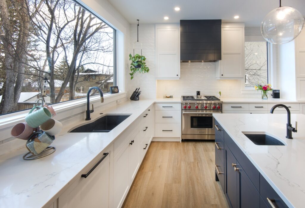 Modern kitchen with white cabinets, black hardware, a large window, marble countertops, a stainless steel stove, a black range hood, an island with a sink, light wood floors, and a hanging glass globe light.