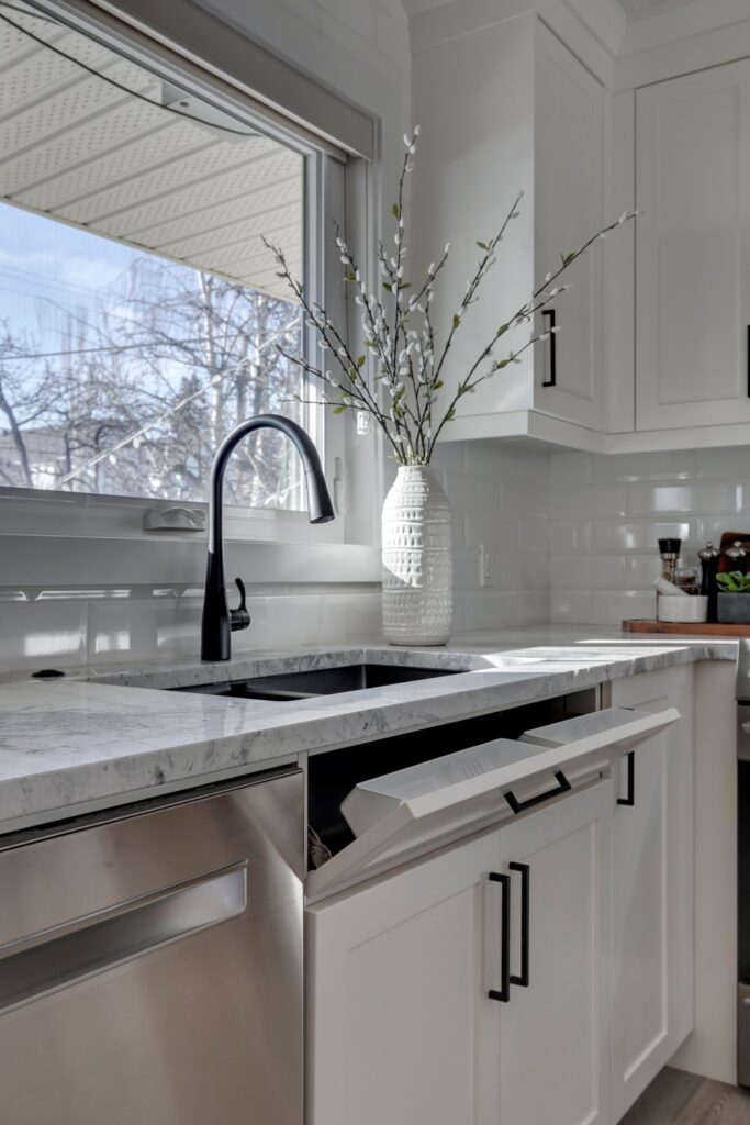 A modern kitchen features a marble countertop with a black faucet, a partially open dishwasher, white cabinets with black handles, and a white vase with tall branches on the counter near a large window.
