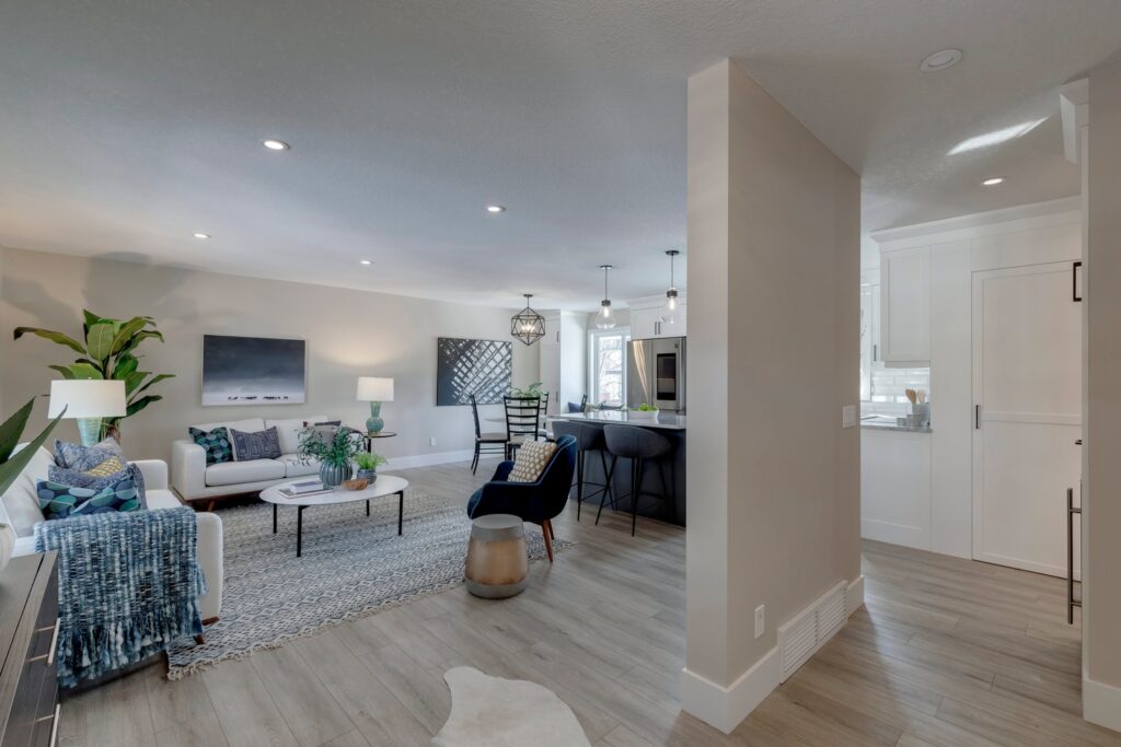Modern open-concept living room and kitchen with neutral walls, wood flooring, stylish furniture, plants, and pendant lights; white sofa, blue and black chairs, and a cozy, inviting ambiance.