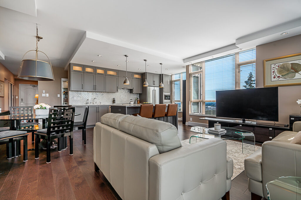 Modern open-concept living room and kitchen with gray cabinets, stainless steel appliances, a large TV, beige leather sofas, a glass dining table, and large windows letting in natural light.