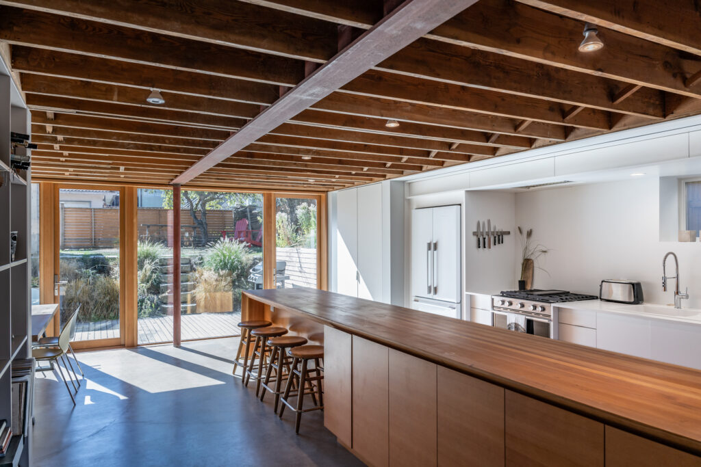 Modern kitchen with exposed wooden ceiling beams, a long wooden island with barstools, white cabinets, and large glass doors opening to a sunlit patio with greenery outside.