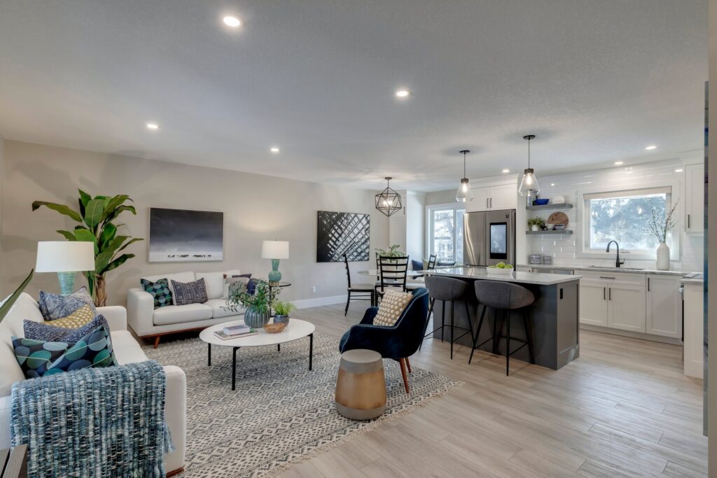 Open-concept living room and kitchen with modern decor, neutral walls, light wood floors, white sofa, blue armchair, indoor plants, round coffee table, bar stools at a kitchen island, and pendant lighting.