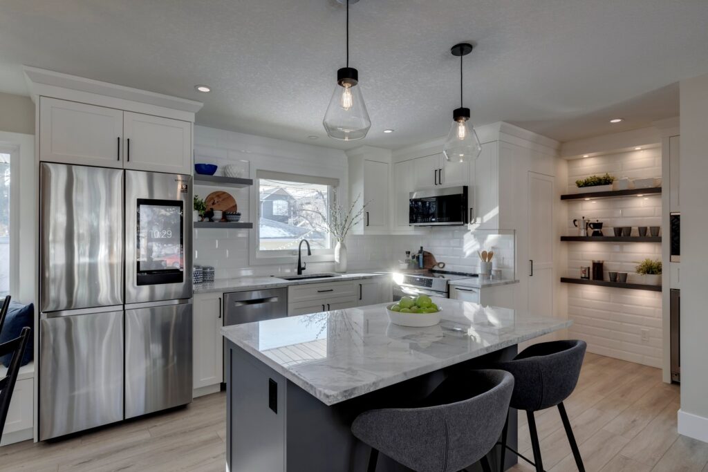 Modern kitchen with white cabinets, stainless steel appliances, a marble island with three gray chairs, pendant lights, and an open pantry with shelves and decor. Sunlight streams in through the window above the sink.