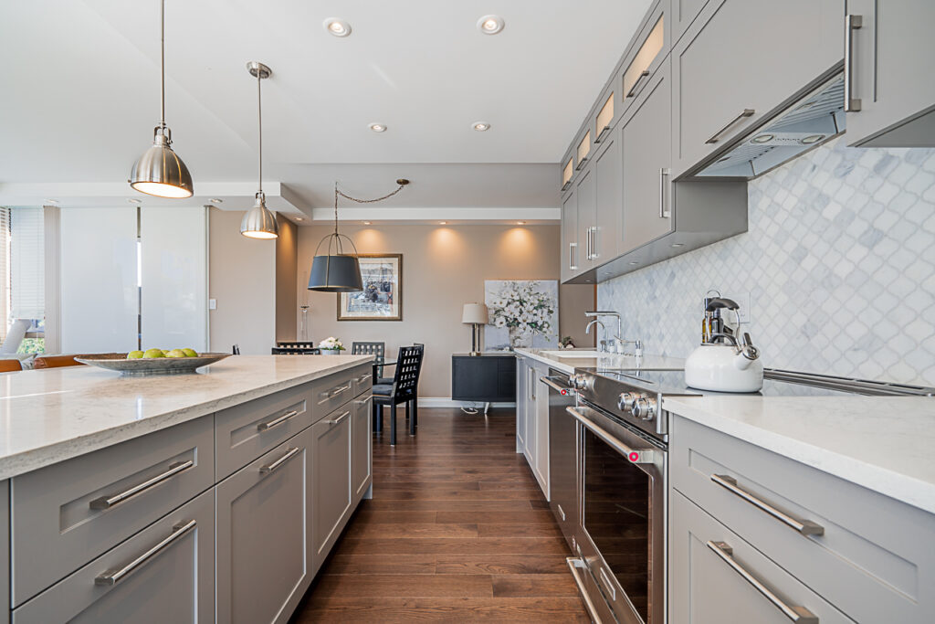 Modern kitchen with gray cabinets, white marble countertops, pendant lights, stainless steel appliances, and a tile backsplash. Dining area with a table and chairs is visible in the background.