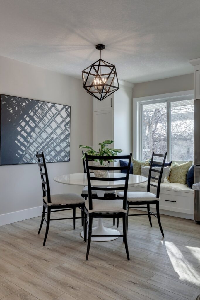 A modern dining area with a round white table, four black chairs, geometric pendant light, abstract wall art, and a window seat with cushions beside a large window. Light wood flooring throughout.