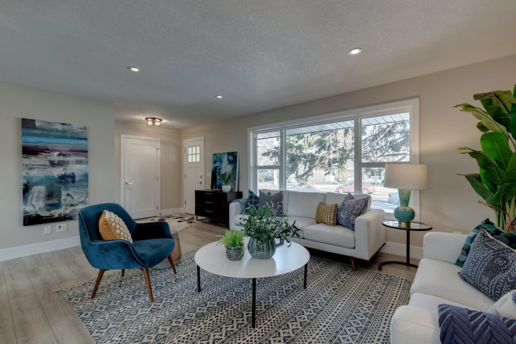 Modern living room with a white couch, blue armchair, patterned rug, round white coffee table with plants, large window letting in natural light, abstract artwork, and a tall potted plant in the corner.