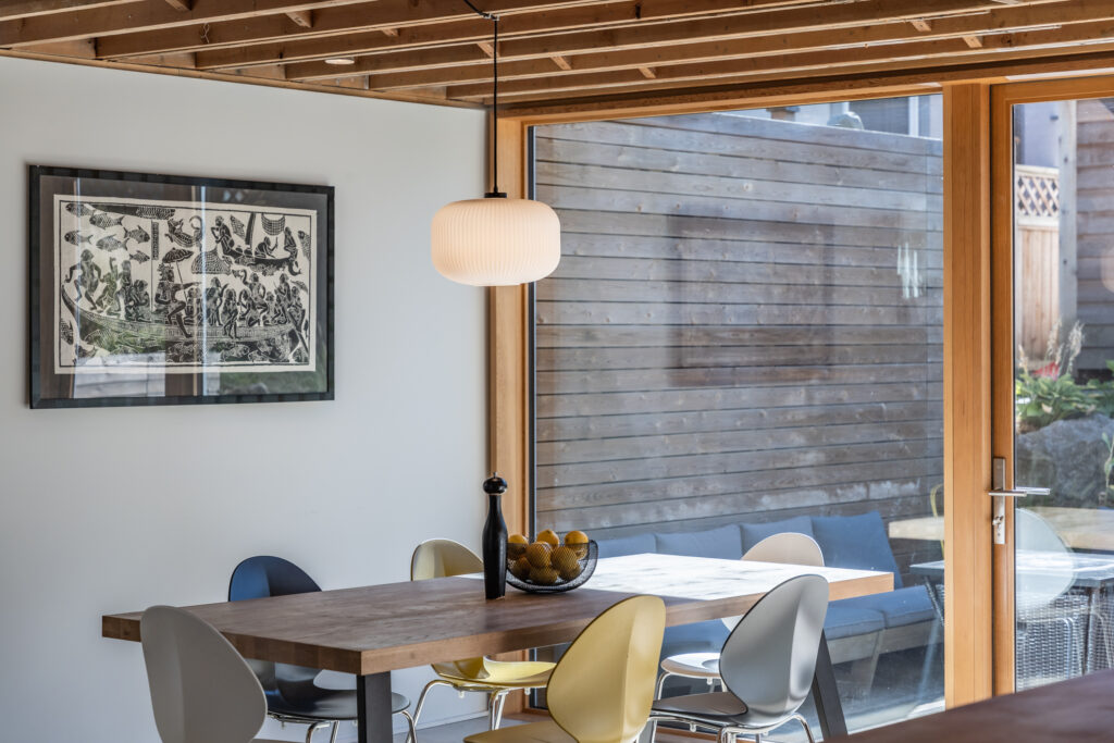 A modern dining area with a wooden table, mixed-color chairs, a bowl of pears, and a pendant lamp. Large glass doors provide natural light, and a framed artwork hangs on the white wall.